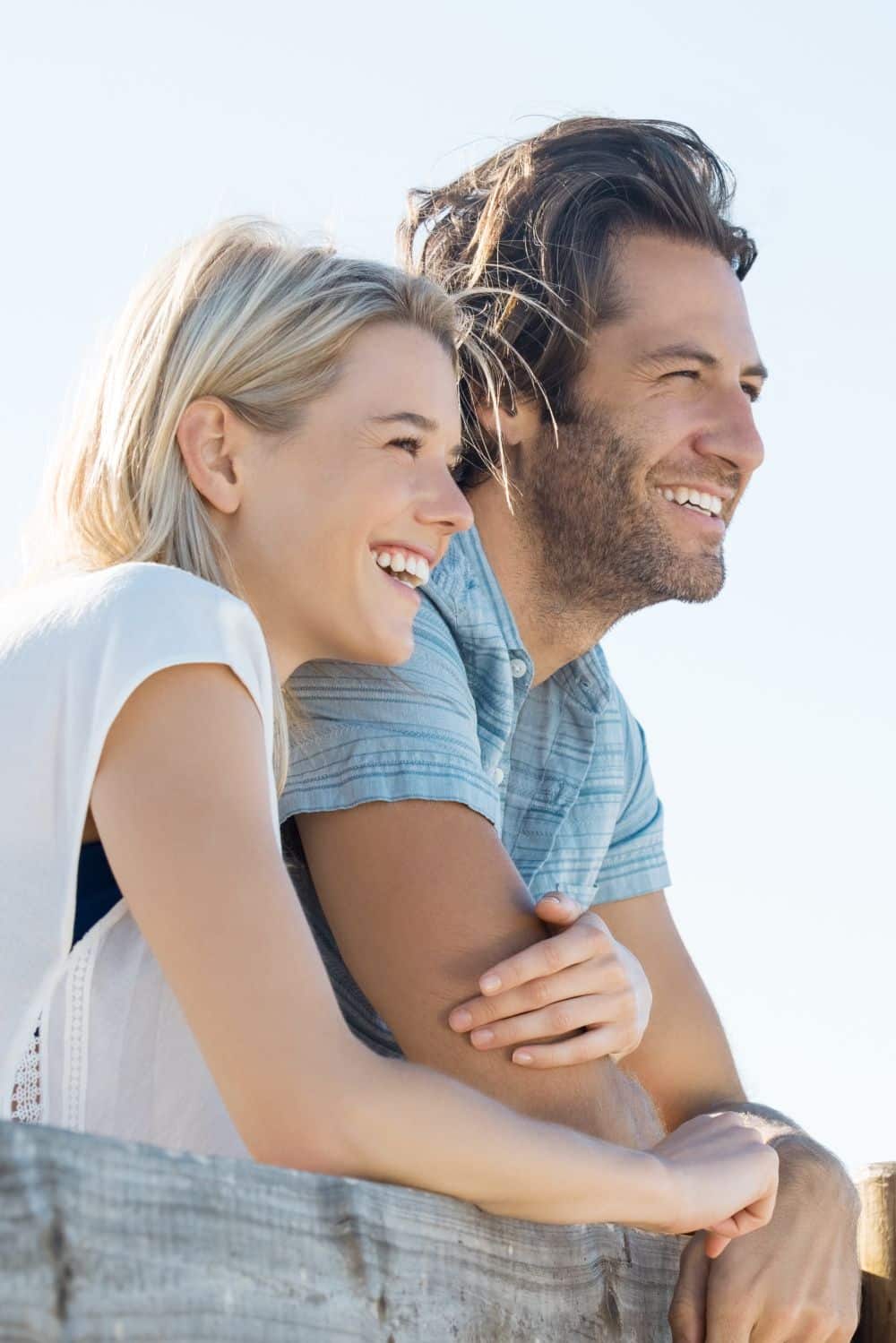 Happy young couple leaning over fence and looking away, copy space. Smiling couple thinking about the future. Young woman embracing her boyfriend outdoor during the sunshine.