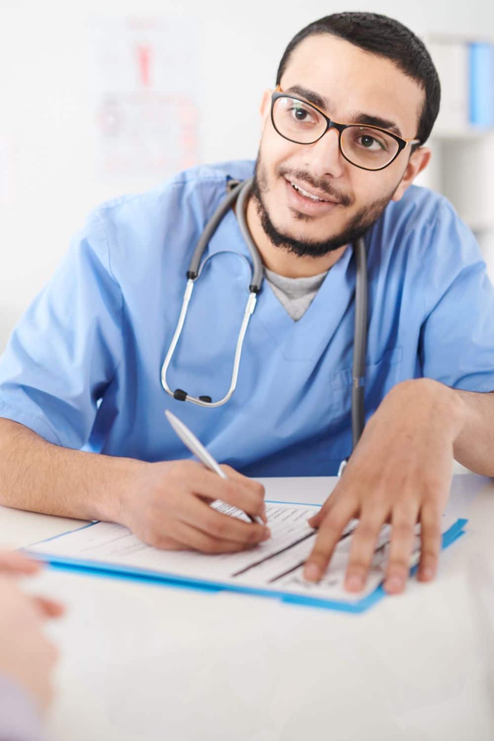 Nurse wearing glasses sitting at desk in office and listening intently to patient while making notes