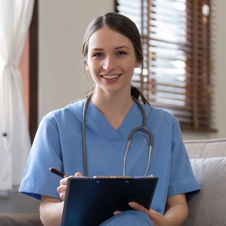 Portrait of young attractive smiling nurse wearing uniform and stethoscope sitting on sofa. Home nurse. Caucasian woman working in nursing home.